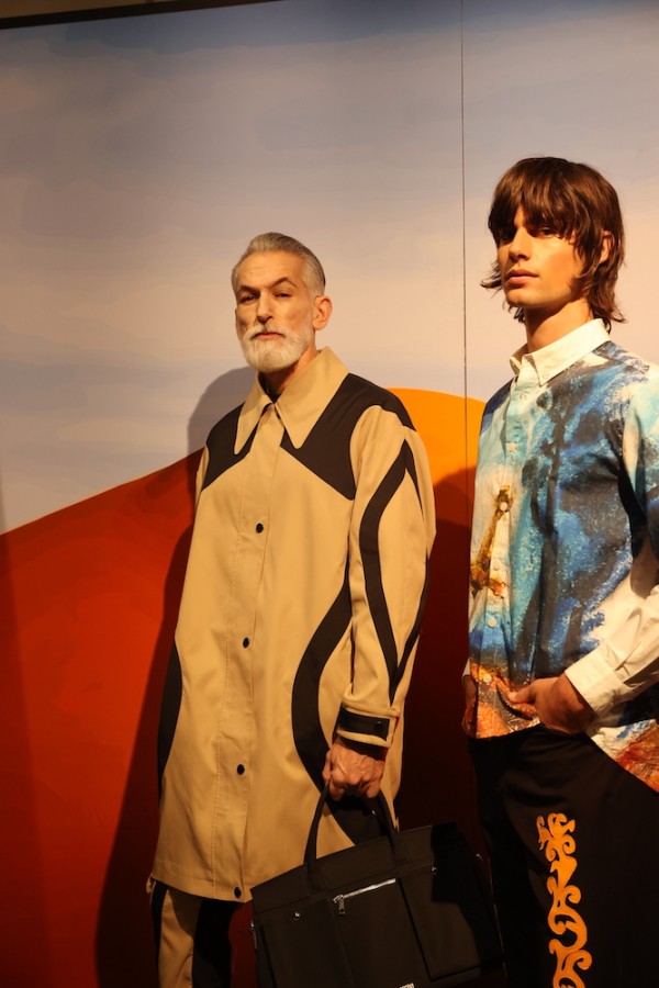 Two models stand in front of a blue and orange backdrop. A model with white facial hair wears a beige jacket and is holding a black handbag on the left; a model wearing a patterned blue shirt and black pants with orange patterns stands on the right.
