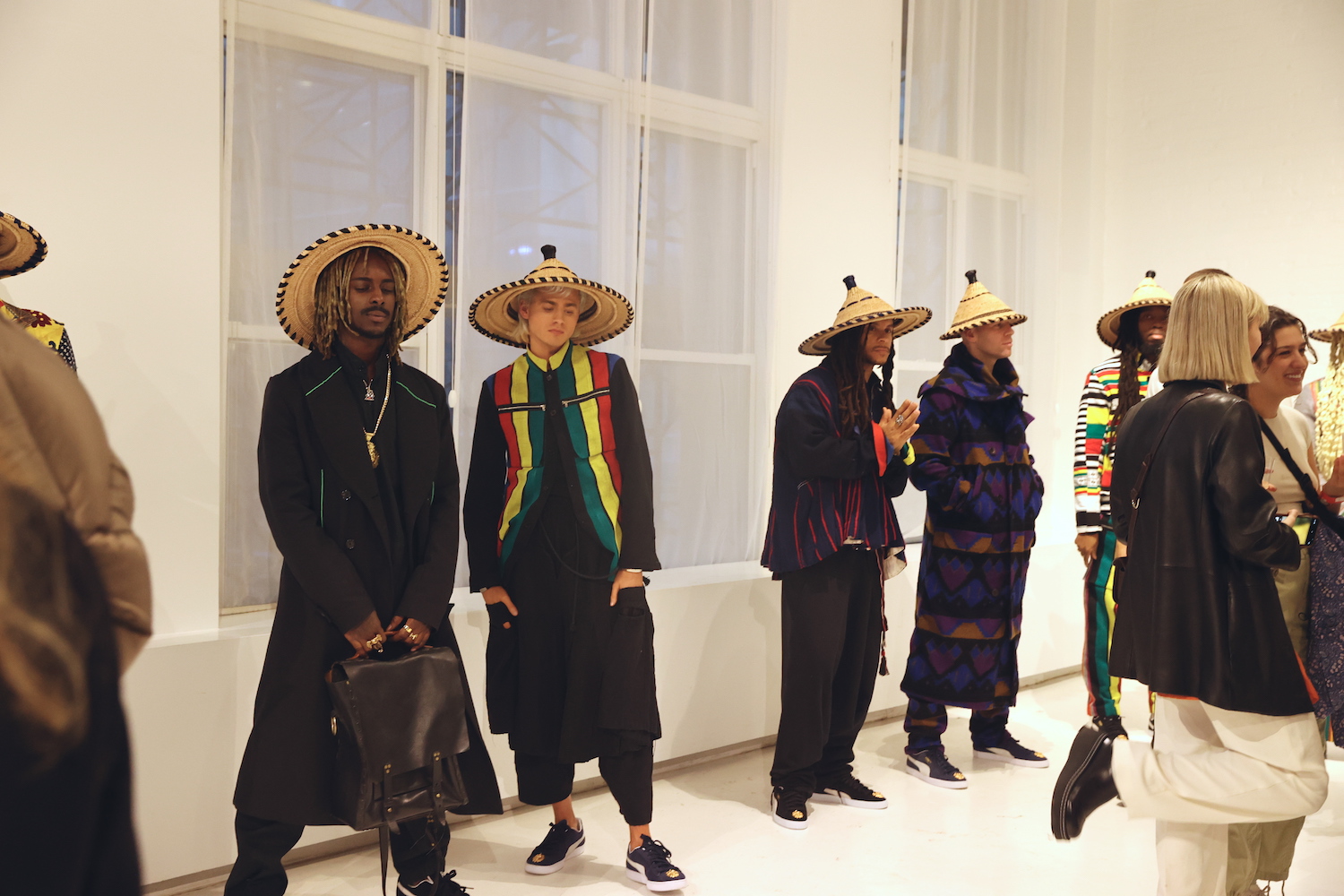 A group of people standing inside a white studio space. Several models wearing straw hats and brightly-colored outfits.