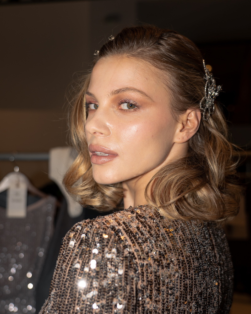 A flash photograph of a model with curled, blonde hair looking at the camera, wearing a sparkly silver top and matching sparkly hair clip.