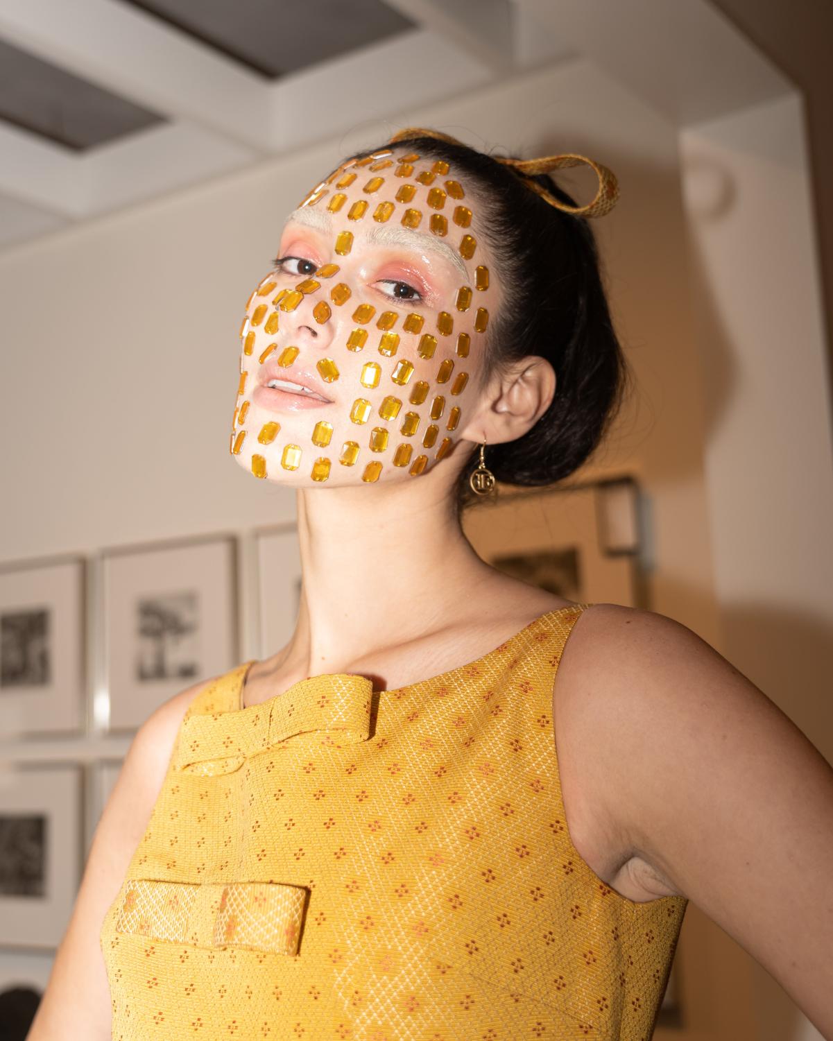 A flash photograph of a female presenting model looking into the camera. Her face is decorated with many yellow gemstones, salmon eyeshadow, and bleached eyebrows. She wears a yellow dress with bows down the middle that match the one on her head.