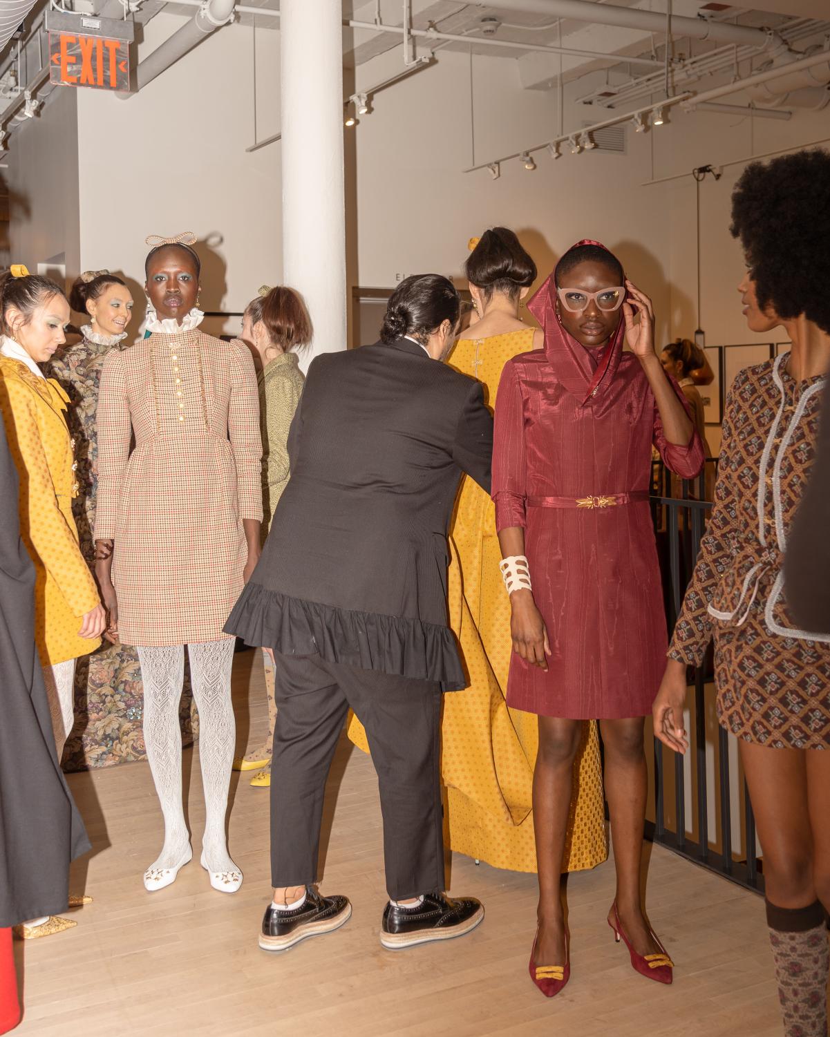 Flash photograph of many female presenting models lining up before walking the runway. Victor, the designer, is pictured in the middle with his back to the camera as he organizes the models.