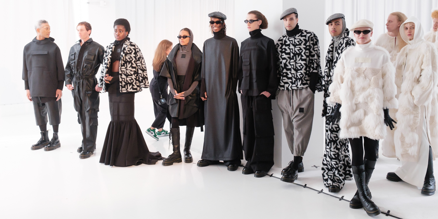 A row of twelve models stand in a line. They wear various black-and-white outfits. The room is completely white and brightly lit.