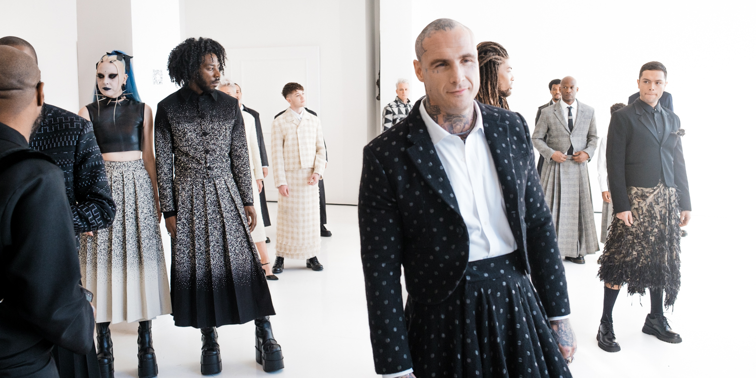 A model stands at the forefront of the image, wearing a white button down shirt, a black blazer with white polka dots, and a black skirt with white polka dots. Behind him are various other models in black-and-white outfits, standing in a white room.