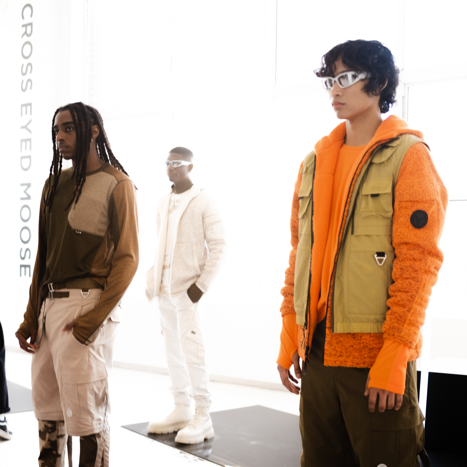 Three models are pictured. The model to the right is wearing an orange jacket, a brown vest, brown pants, brown boots and white sunglasses. The model to the left is wearing a brown shirt, brown and beige pants and brown shoes. The model in the middle is wearing all white — a shirt, a jacket, pants, boots and sunglasses.