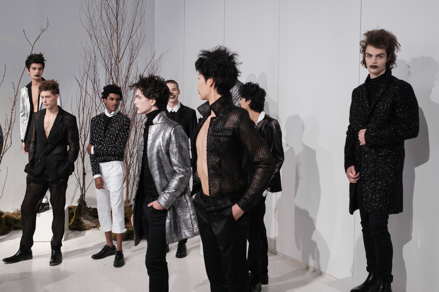 A group of nine models wear various black, white and silver outfits. In the background are leafless, brown trees.