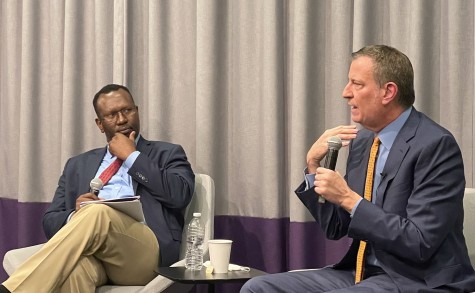 Bill de Blasio scowls while wearing a blue shirt, blazer, dark pants and a yellow tie. He sits while speaking into a microphone. Next to him is a man, also sitting and holding a microphone, in a blue shirt, blazer, khakis and a red tie. They are in front of a white curtain with a purple stripe at the bottom.