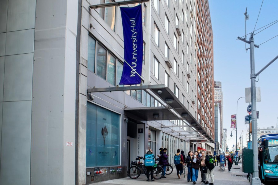 The exterior of N.Y.U.s University Hall, with a purple banner. People gather outside and an M.T.A bus is pictured on the street in front of the building.