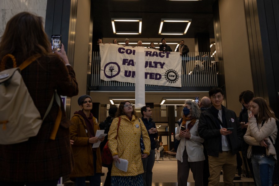 Ten people stand in front of a large white banner being dropped from a balcony above them which reads “Contract Faculty United” in purple with a circular logo reading “U.A.W.” and an N.Y.U. torch. Seven people are holding the banner and a long single piece of paper covered in text.