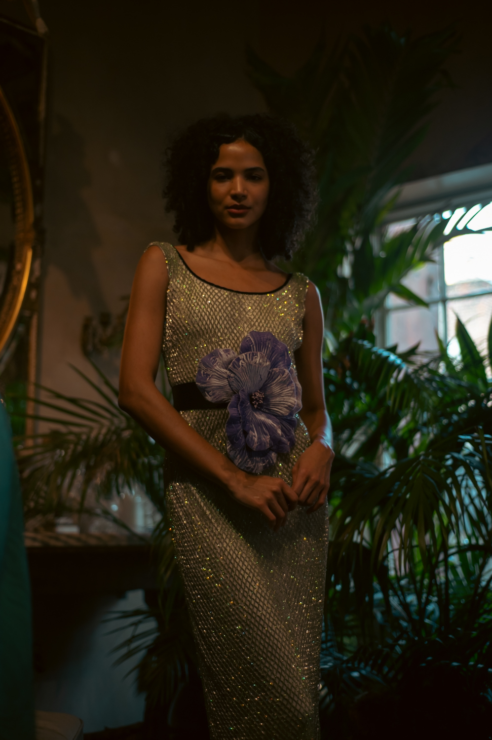 A model wearing a white, fishnet-patterned dress with green sequins, holding a purple flower.