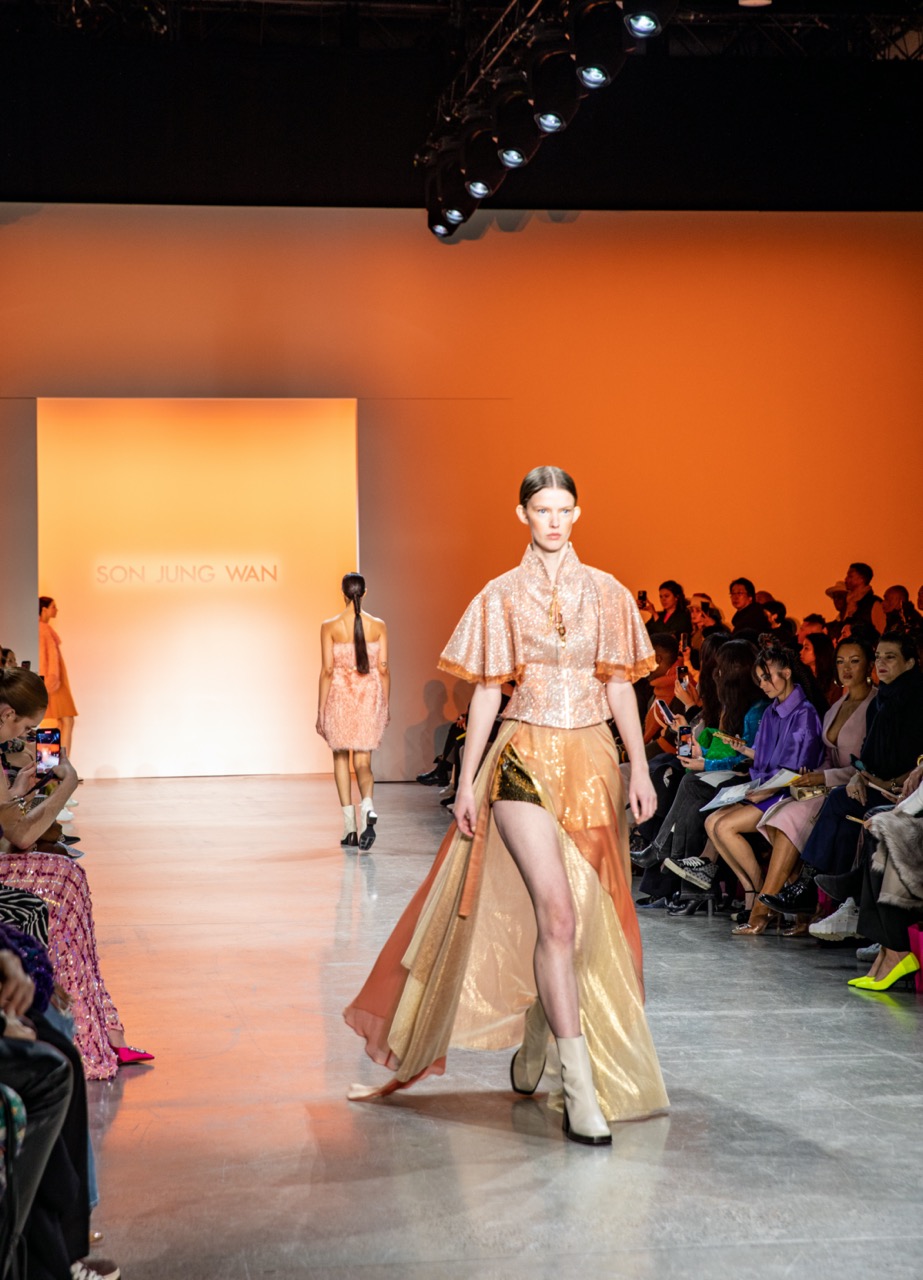 A model walking down a runway lit with orange light, wearing a sparkly orange top, a long sheer orange skirt with a slit, cream-colored boots and slicked-back hair.