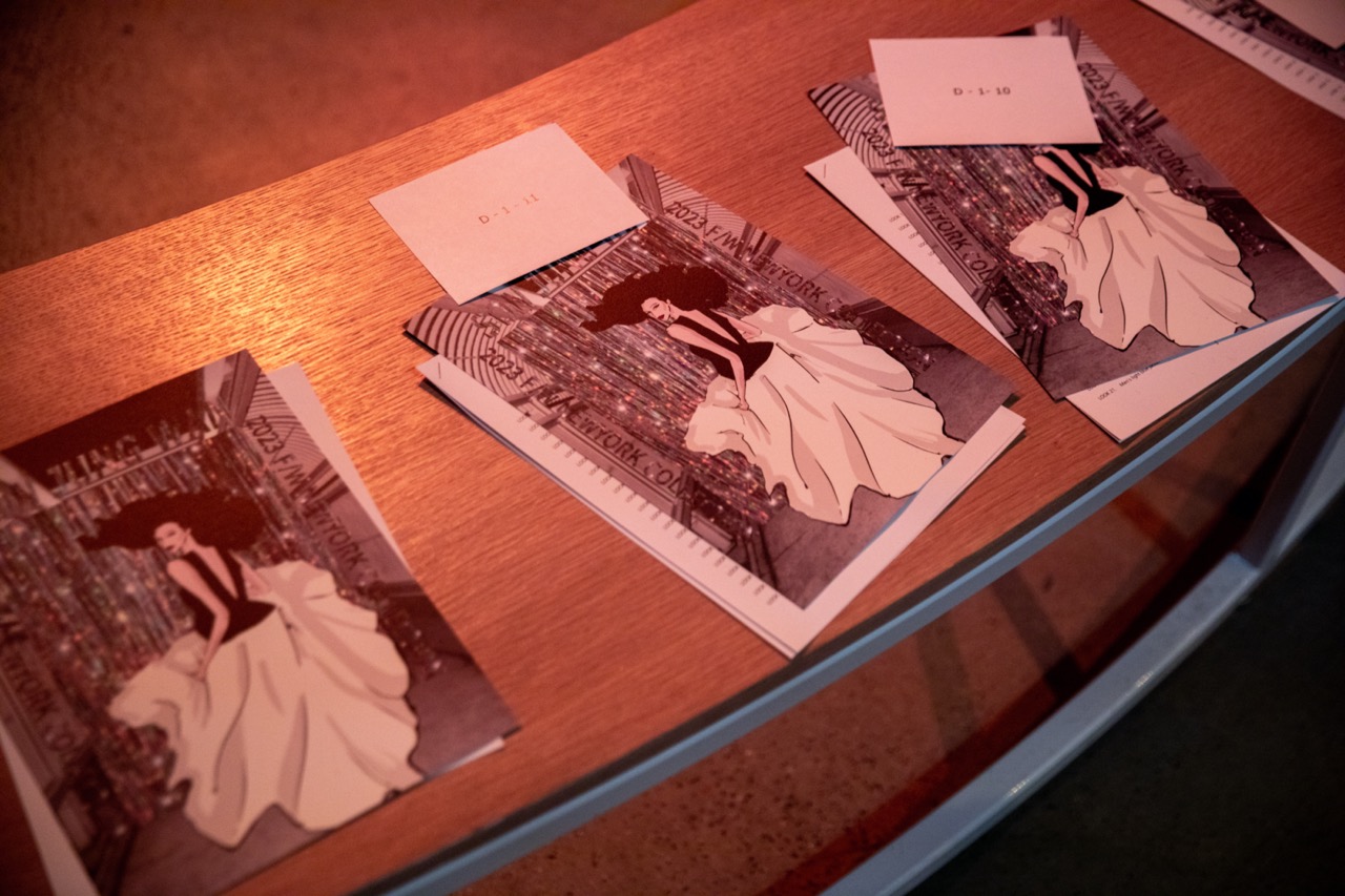 A photo of a wooden table with three cards on top. Each card features an illustration of a model with long, curly black hair walking down a runway, looking back at the camera and wearing a long flowy white skirt and a black top.