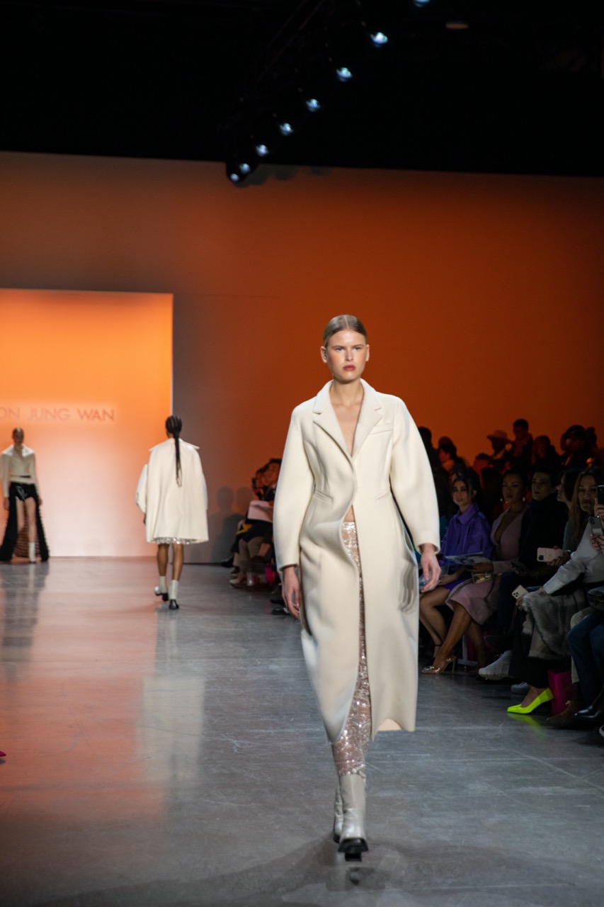 A model walking down a runway lit with orange light, wearing a form-fitting white coat, a sparkly long skirt, cream boots and slicked-back hair.
