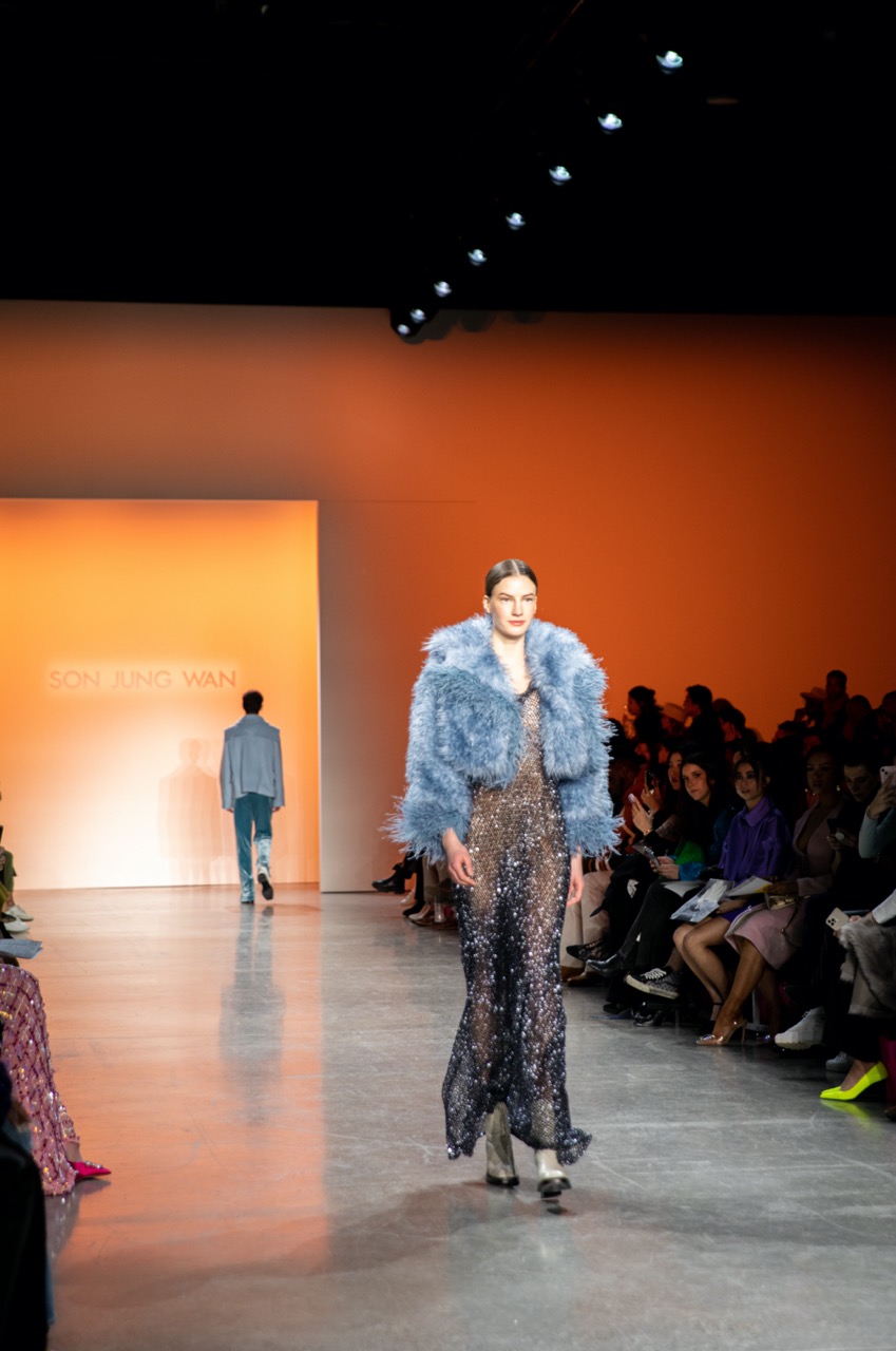 A model walking down a runway lit with orange light, wearing a fluffy blue jacket, a sparkly black long dress, cream boots and slicked-back hair.