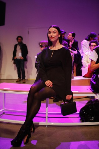 Grace Caroline Currey poses while sitting in the audience. She wears a black dress and black high heels, and carries a black handbag.