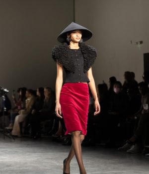 A model walks down the runway sporting a black conical hat, a red velvet skirt, and a black blouse with structured, feathered sleeves.