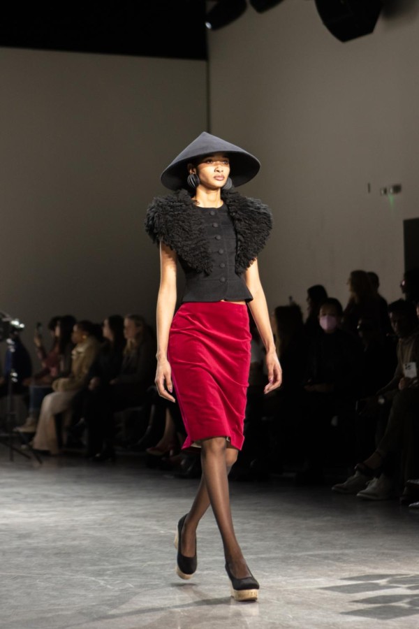 A model walks down the runway sporting a black conical hat, a red velvet skirt, and a black blouse with structured, feathered sleeves.