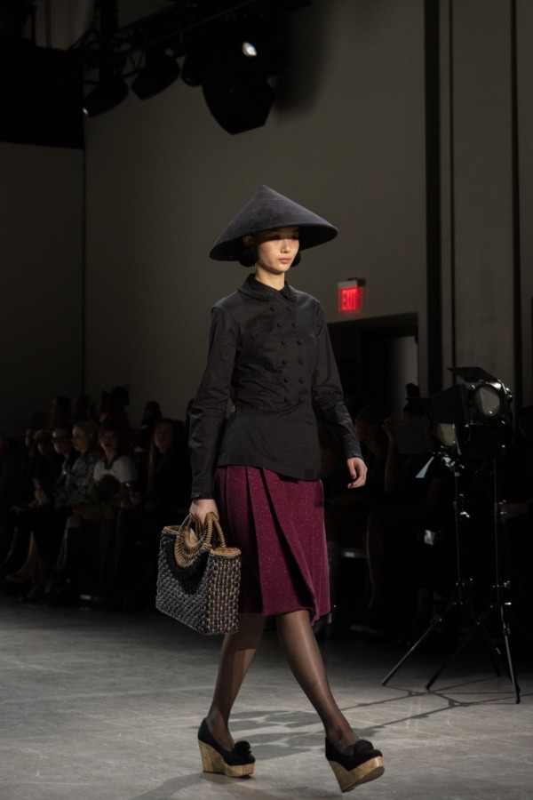 A model walks down the runway sporting a black conical hat, a black dress shirt and a maroon skirt. They carry a woven handbag.