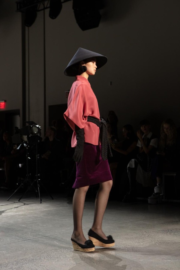 A model walks down the runway sporting a black conical hat, a salmon-colored shirt, and black, elbow-length gloves. Around her waist is a black, woven cord that sits casually above a purple velvet skirt.
