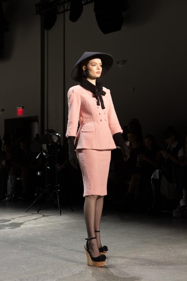 A model walks down the runway sporting a black conical hat with a pink coat and skirt. She wears black, elbow-length gloves that pair with the black ruff around her neck.