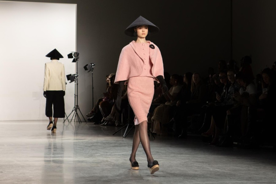 A model walks down the runway sporting a black conical hat, a pink coat and a skirt. She wears black, elbow-length gloves that pair with her black spherical earrings and brooch.