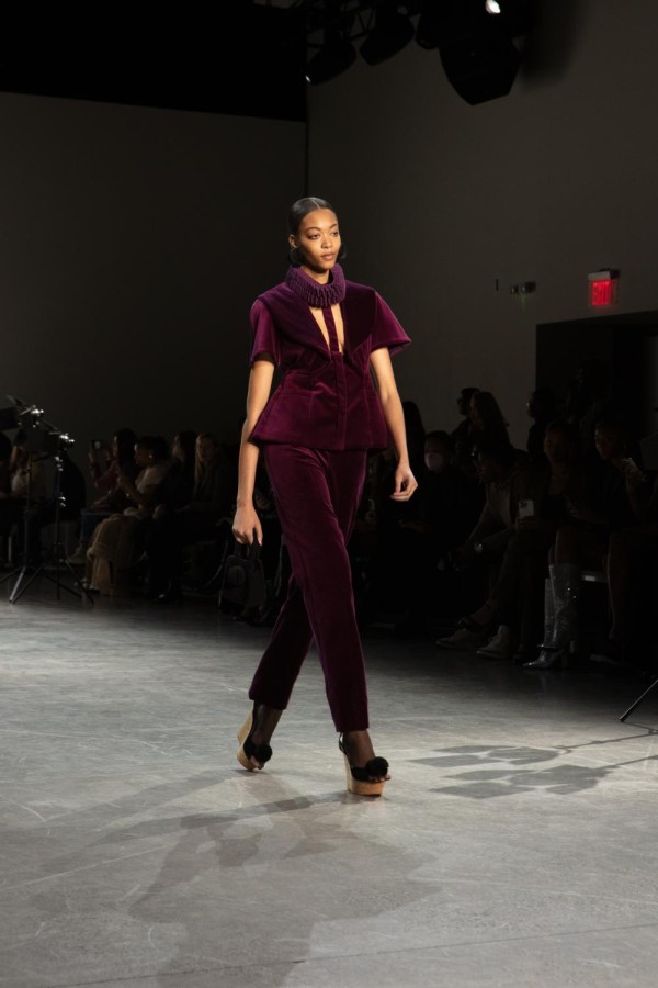 A model walks down the runway sporting purple velvet outerwear and a purple ruff around her neck. She wears black, elbow-length gloves that pair with her black spherical earrings.