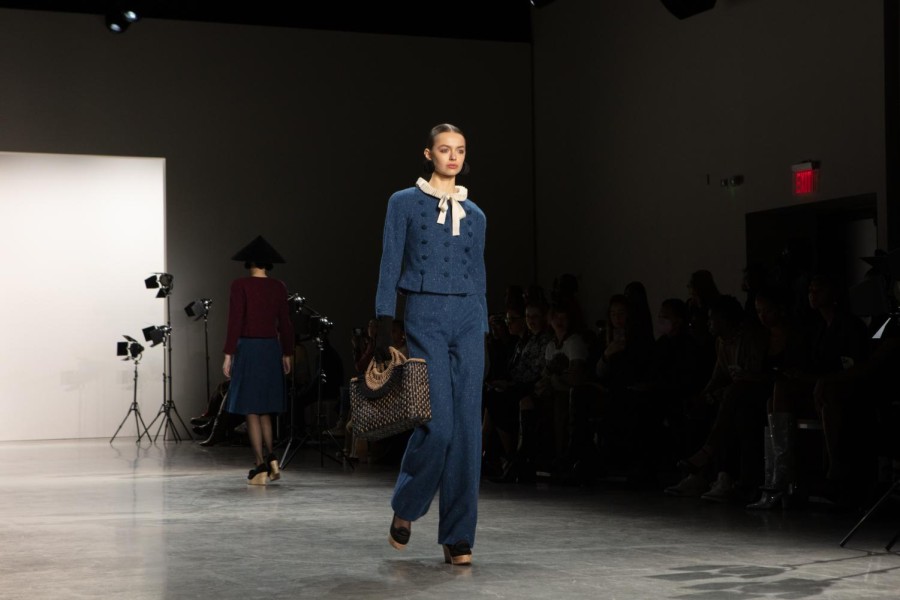 A model walks down the runway sporting a blue coat and slacks with a white neck ruff. She wears black spherical earrings and black elbow-length gloves, and is carrying a woven handbag.