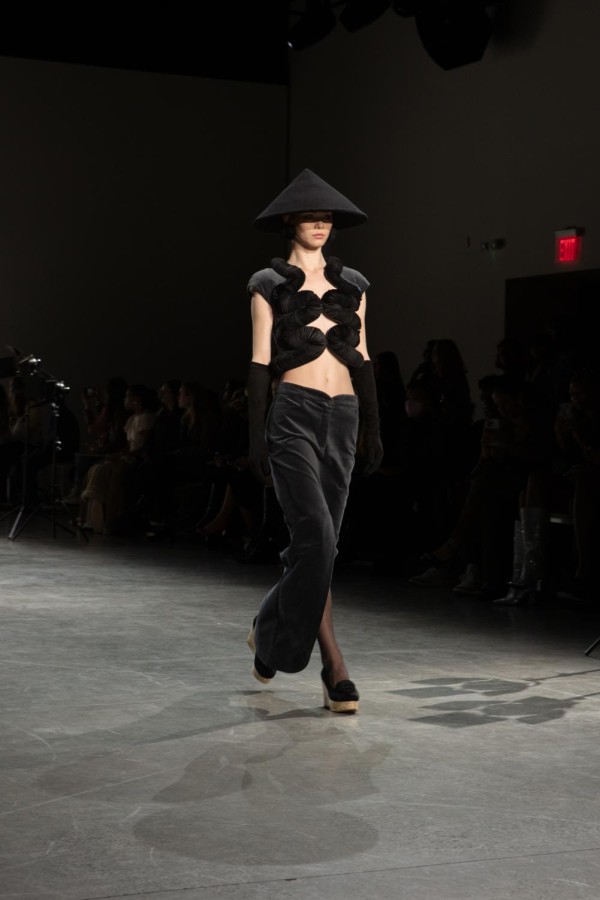 A model walks down the runway sporting a black conical hat with a fur shirt and black velvet pants. She wears black spherical earrings and black, elbow-length gloves.
