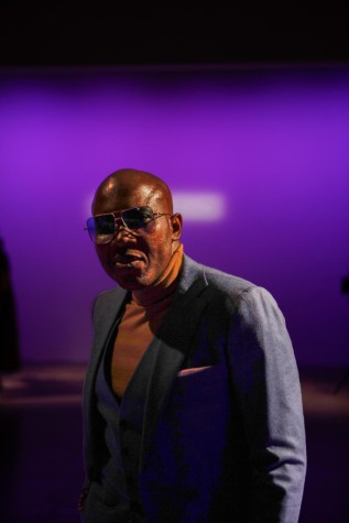 Taiwo Meghoma poses on the runway for the Chocheng fashion show. He wears a blue suit with a yellow turtleneck sweater and sunglasses.