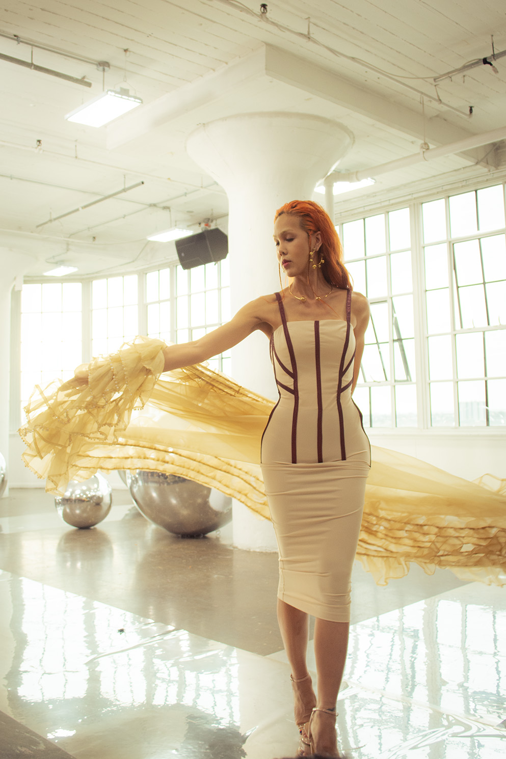 A woman spreads a yellow tulle throw behind her. She is wearing a beige dress with brown accents, and she has orange hair.