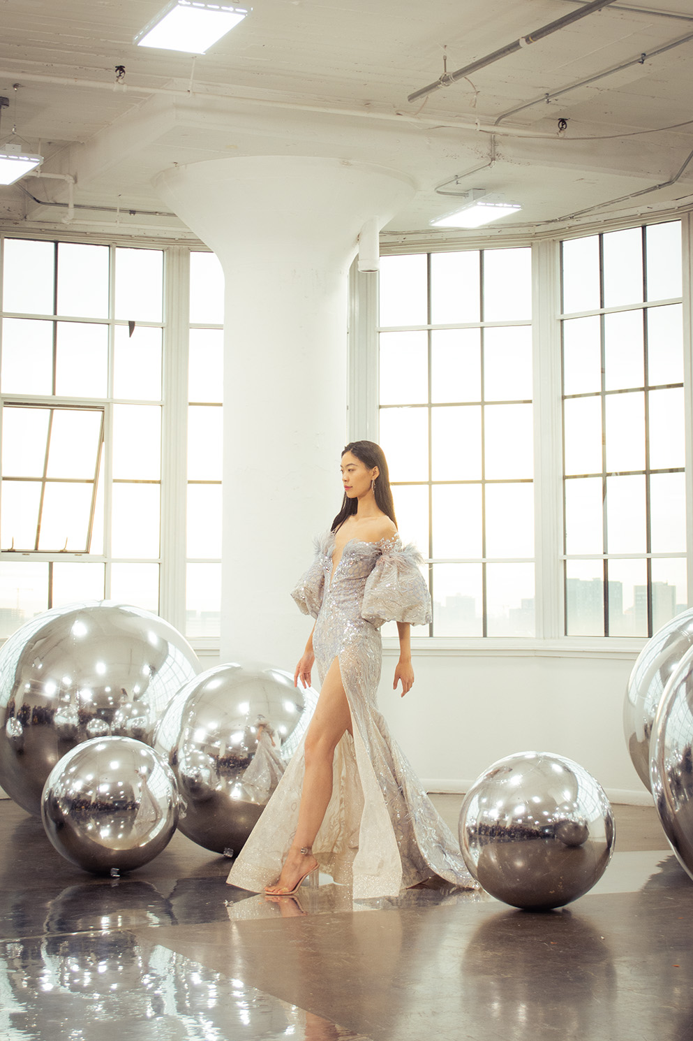 A model wearing a silver maxi dress with puffed sleeves walks between metallic spheres on the ground.