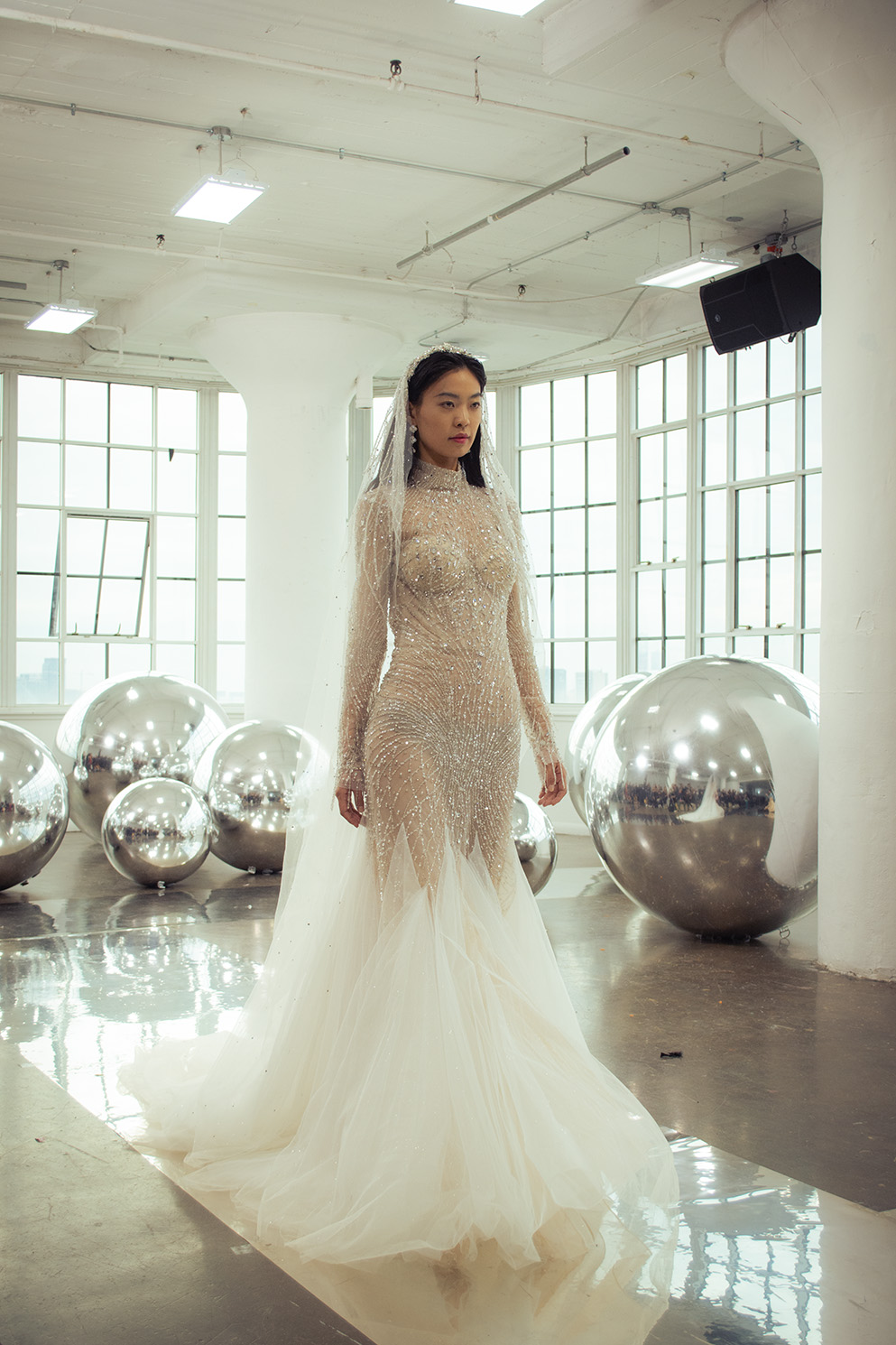 A woman wears a white beaded dress with a matching veil. A window with metallic spheres on the ground is in the background.