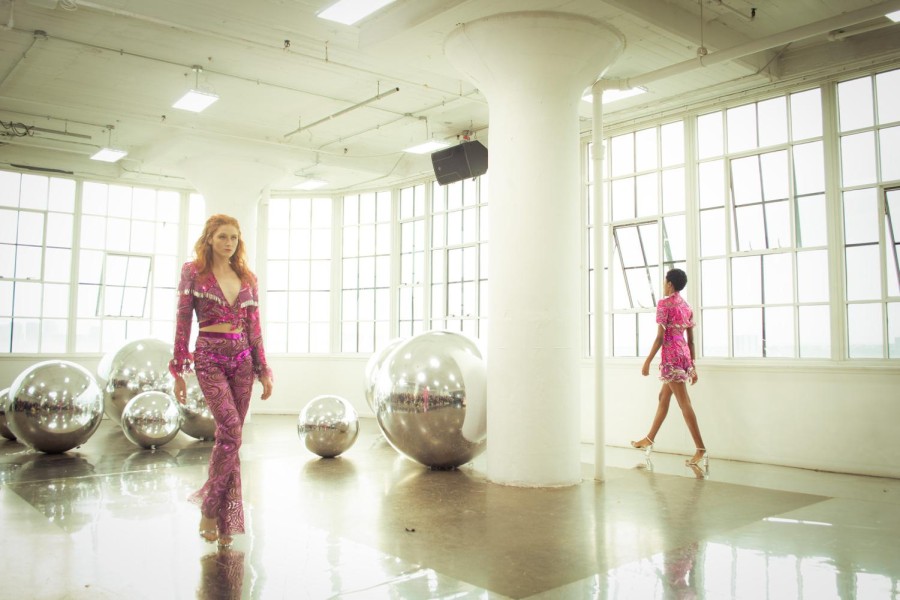 One model wears magenta, sequined pants and a collared shirt, and another wears a magenta, sequined dress. They walk opposite one another, going in opposite directions.