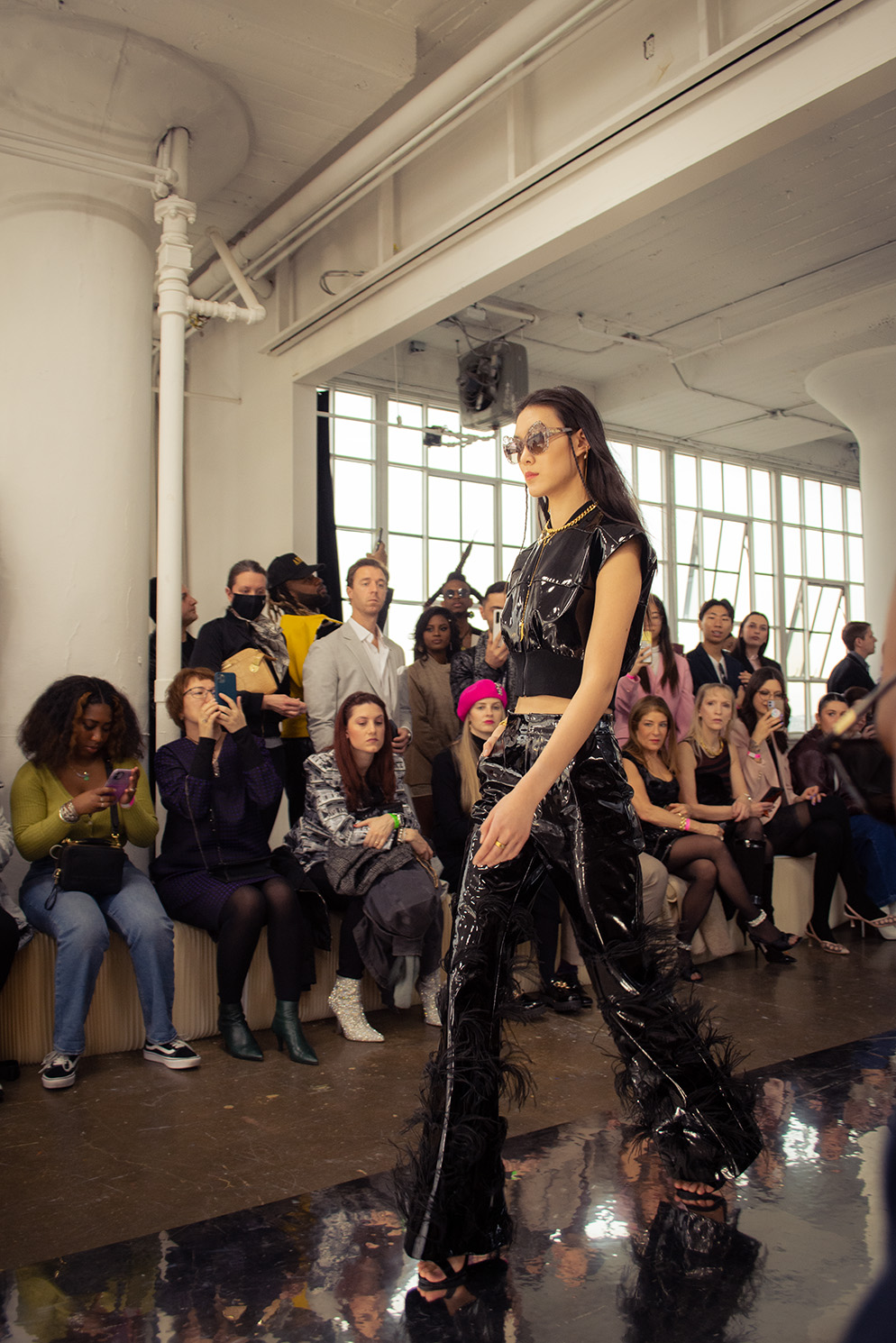 A woman with sunglasses and black, shiny patent leather or vinyl clothing walks forward, with a crowd behind her.