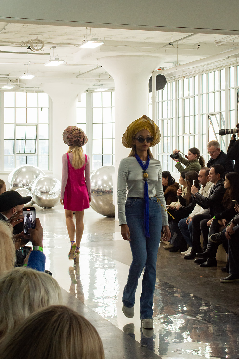 Two models walk, one going toward the back, wearing a hot pink, satin dress with a hat,and the other wearing a satin, long-sleeved top, jeans, a yellow hat and a blue necklace with a gold bead in the center.