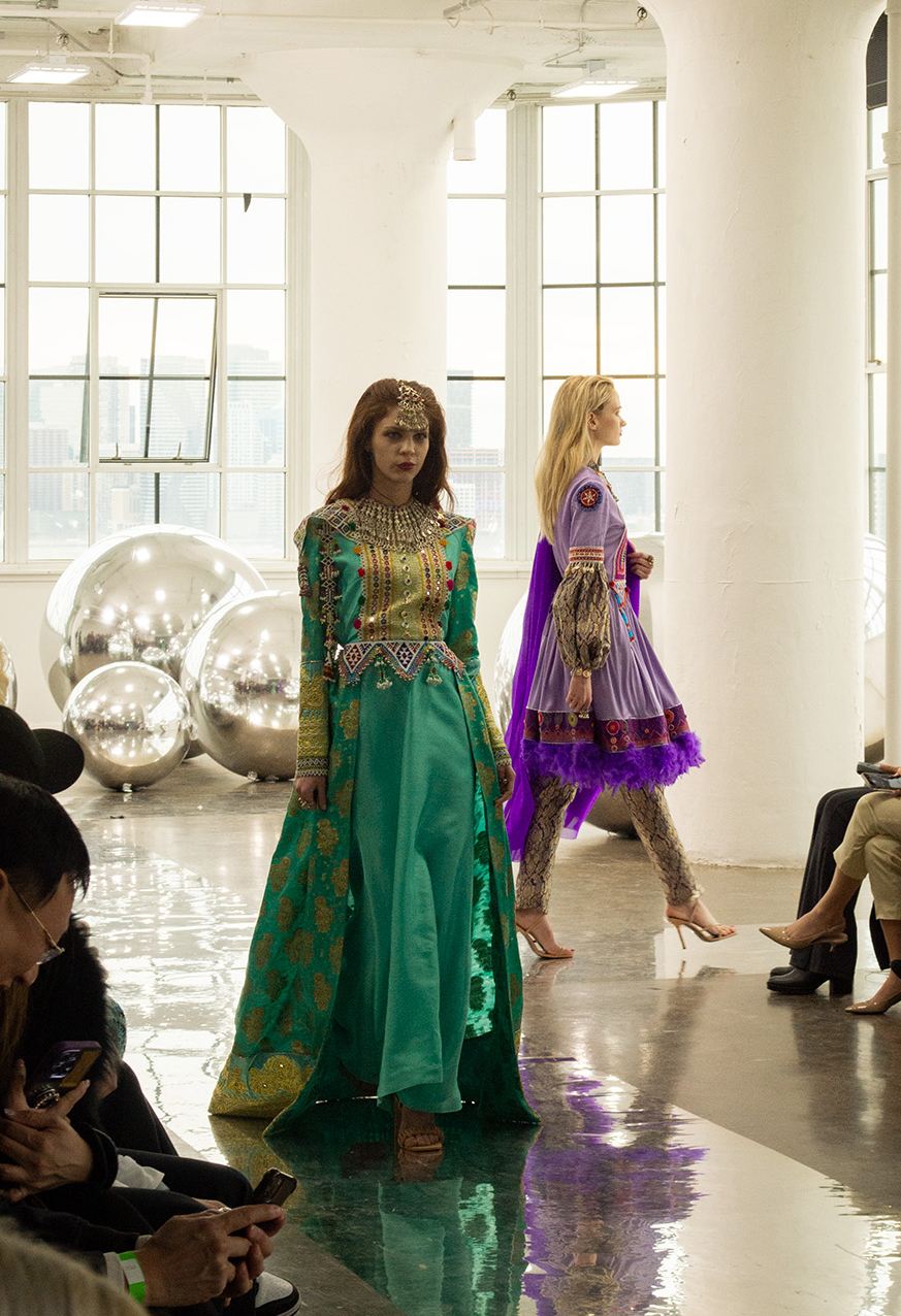 A model walks toward the camera wearing a green dress with an ornate and intricate gold design, as well as a gold head piece. The other model walks to the right behind the first model, wearing a purple dress with similar ornate and intricate designs.
