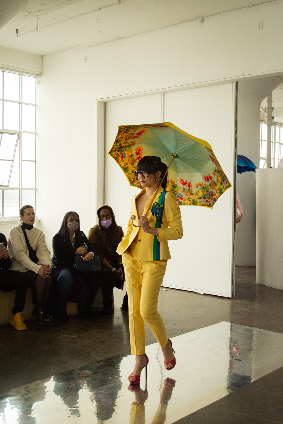 A model walks forward, wearing a yellow suit with a green and blue stripe and a patch shaped like a parrot. She holds a yellow umbrella with flowers painted on the inside.