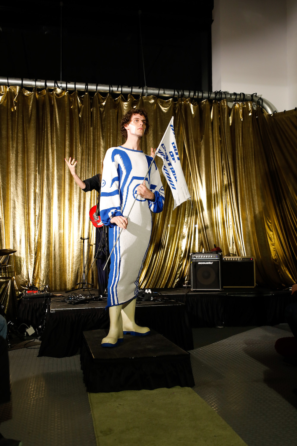 Model dressed in a blue and white long dress with yellow puddle boots holding a matching flag by designers Paradoxvestedrelics, Yxen, and Kids Destroy.