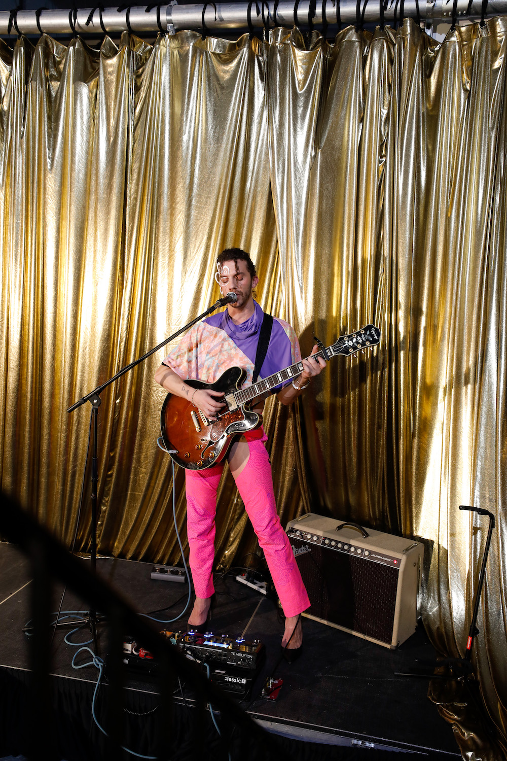 Singer and guitarist Leone performing on stage dressed in a half purple, half multicolored top and bright pink pants by designer Cilium.