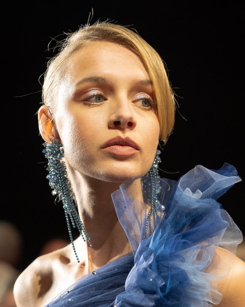 A model posing on the runway, wearing a ruffled blue dress and long, sparkly, blue earrings.