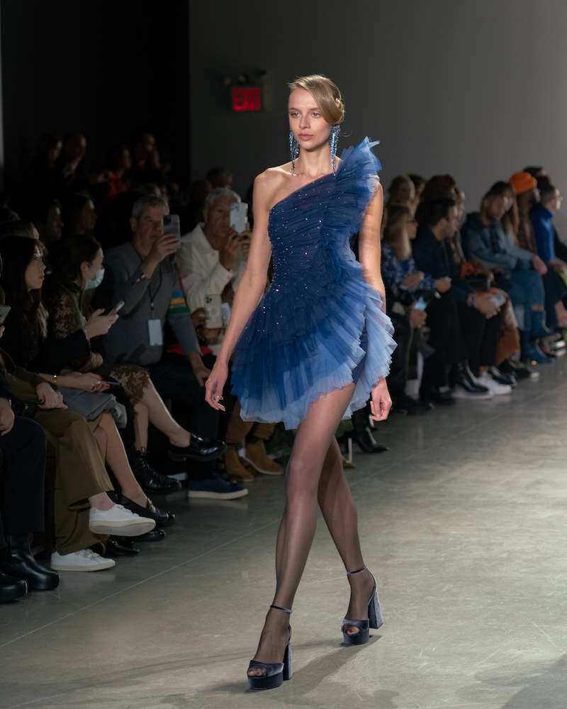 A model walking down the runway wearing a sparkly, ruffled blue dress, long dangly blue earrings and dark blue heels.