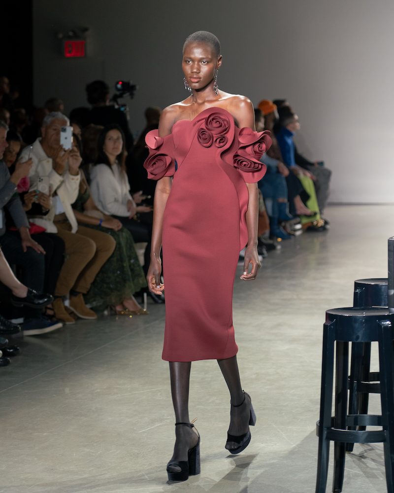 A model walking down the runway wearing a silky, maroon dress with a floral pattern and black heels.