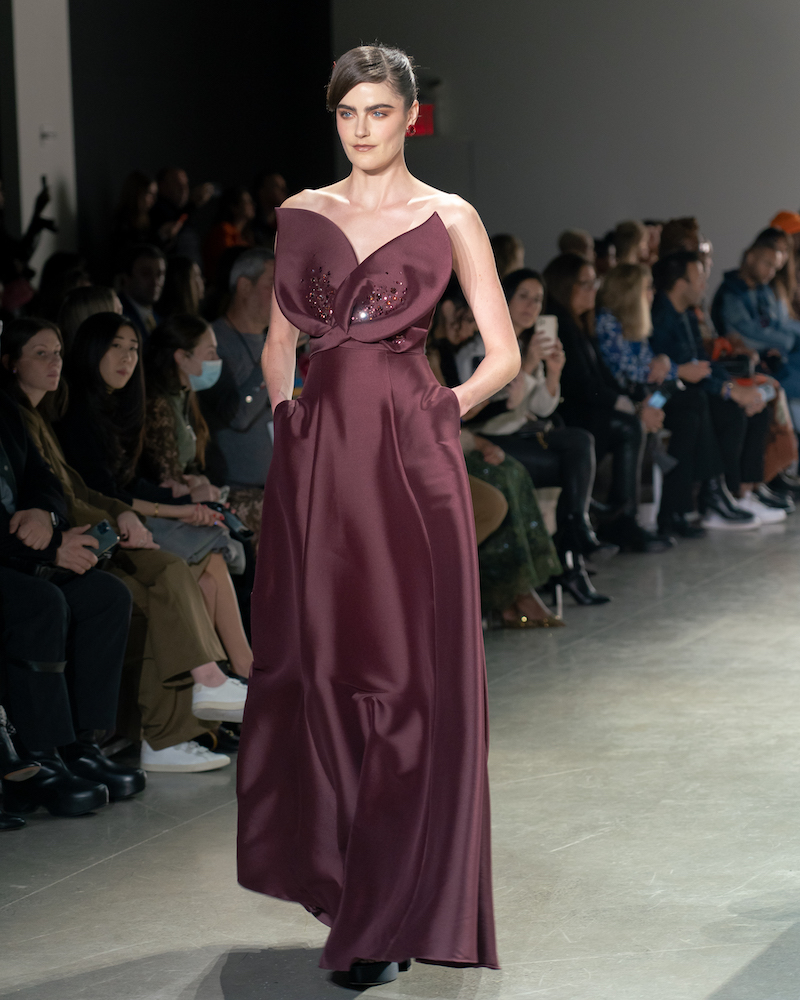 A model walking down the runway wearing a maroon dress with a protruding leaf-shaped detail at the top and black heels.