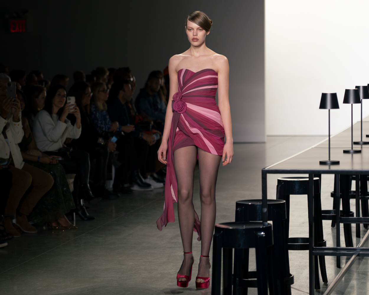 A model walking down the runway wearing a red, pink and maroon mini dress with red heels.