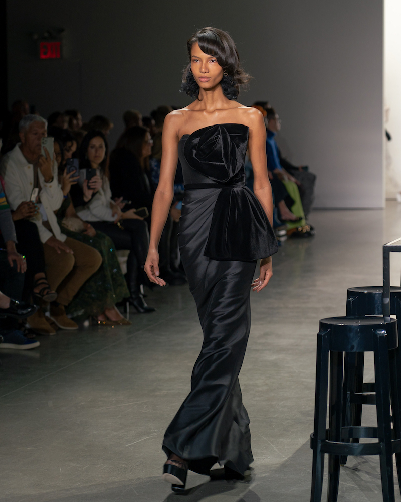 A model walking down the runway wearing a form-fitting black dress with a bow-shaped statement tie around the belt and black shoes.