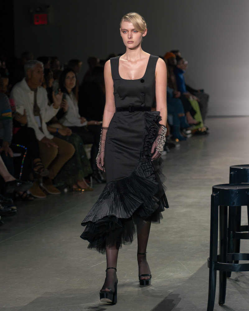 A model walking down the runway wearing a black dress with ruffles at the bottom, black and silver sparkly gloves and black heels.