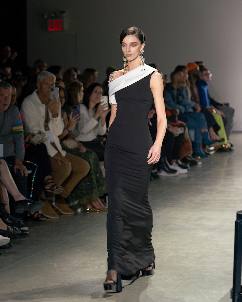 A model walking down the runway wearing a long black dress with a white stripe at the top and black heels.