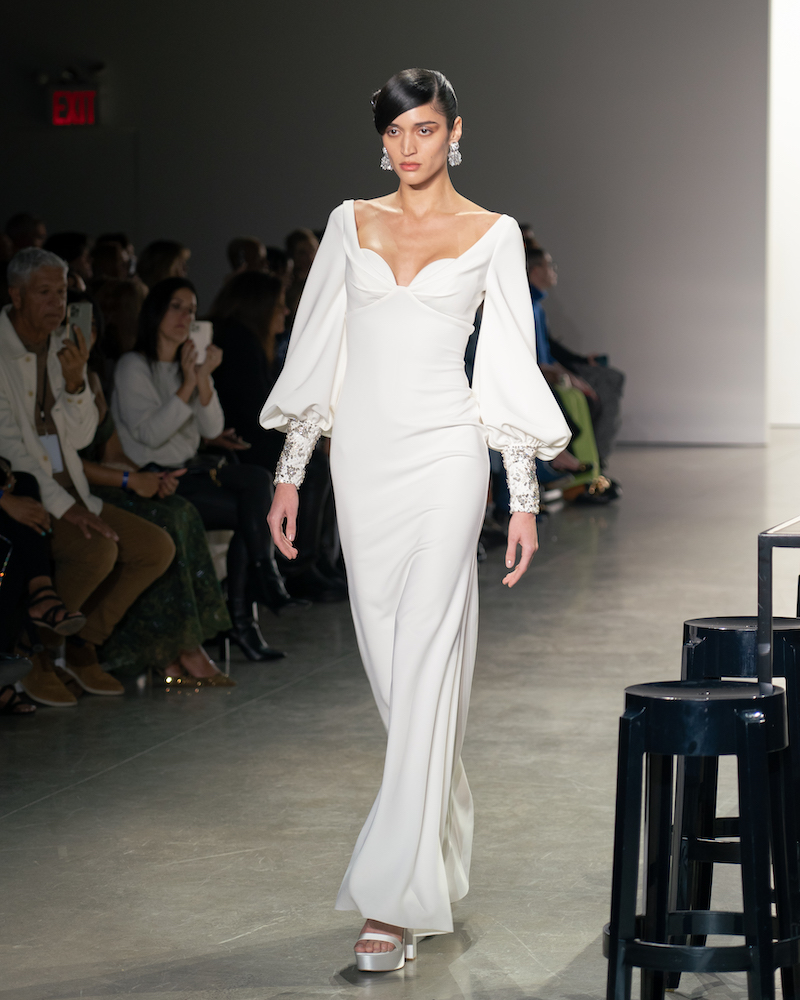 A model walking down the runway wearing a flowy white dress with balloon sleeves and silver heels.