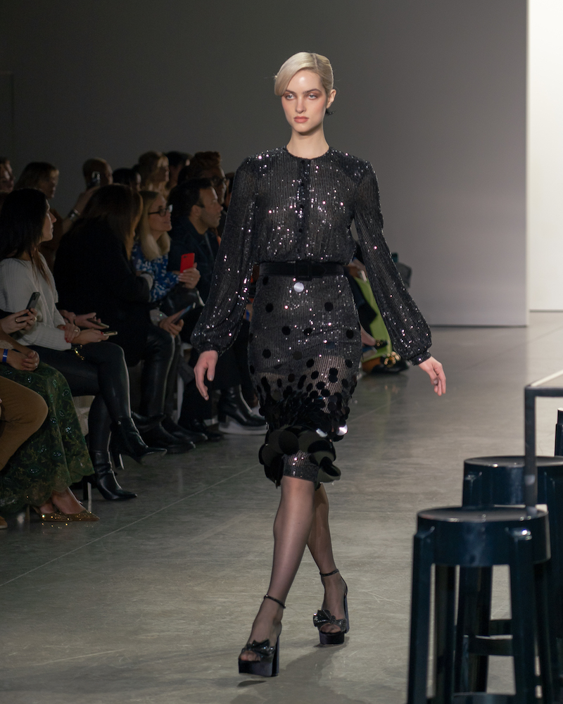 A model walking down the runway wearing a sparkly long-sleeved black dress and black heels.