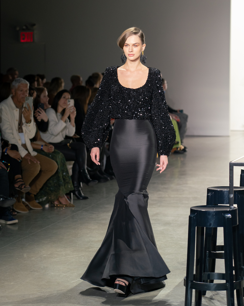 A model walking down the runway wearing a sparkly black top with balloon sleeves, a long, form-fitting silky black skirt, and black heels.