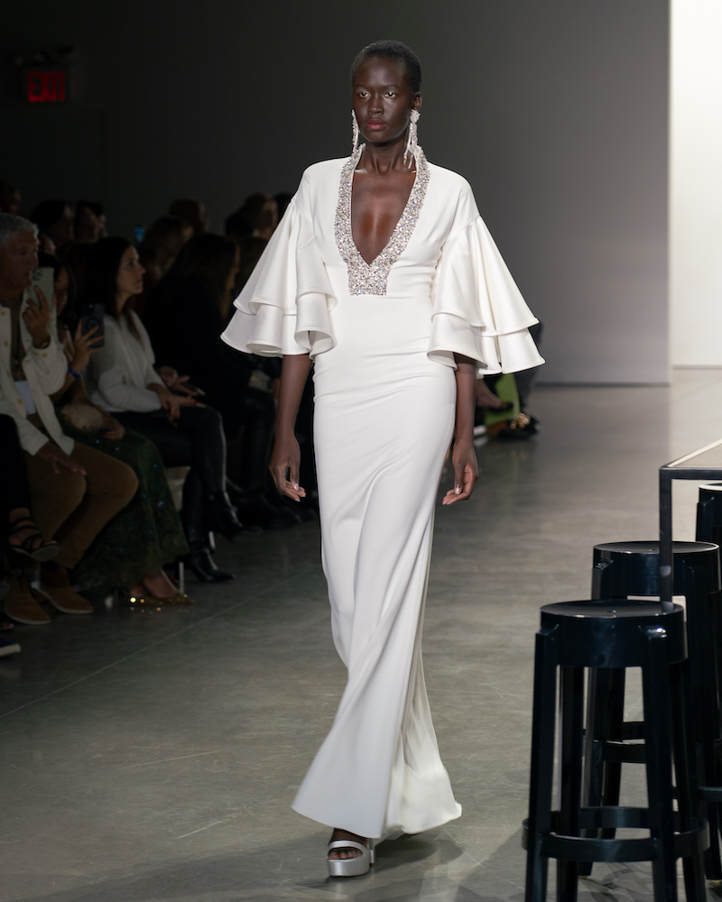 A model walking down the runway wearing a long, white dress with a sparkly collar and layered short sleeves and silver heels.